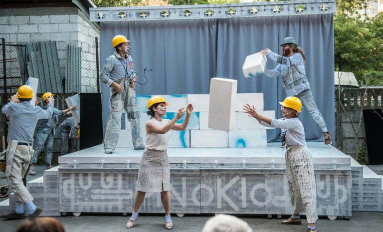 Zdjęcie ze spektaklu Pinokio. Bohaterowie spektaklu w żółtych kaskach budowlanych przerzucają do siebie białe cegły. Z tyłu widać, że z tych cegieł powstaje ściana.