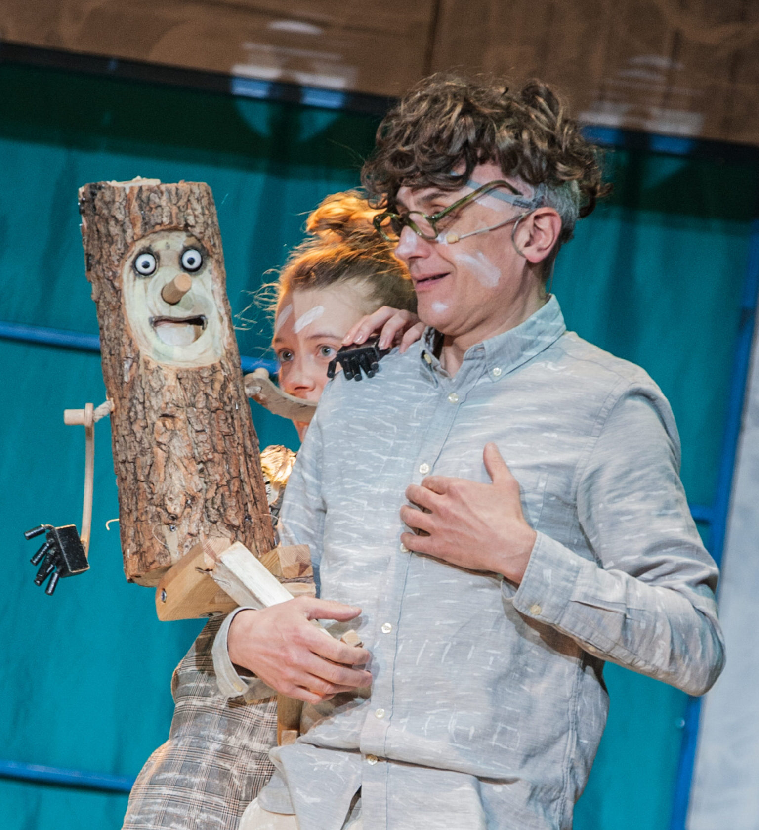 Zdjęcie ze spektaklu Pinokio. Na niebieskim tle dwie posatcie wpatrzone w drewnianą lalkę - Pinokia. Mężczyzna wygląda na wzruszonego i trzyma się za serce.