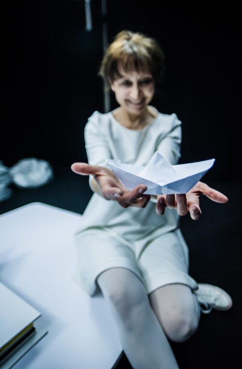Photo from the play The Little Book. In the photo, the smiling actress, dressed in white, holds a paper ship in her outstretched hands.
