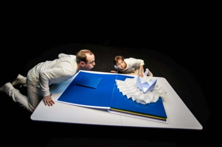 Photo from the play The Little Book. In the photo, a large book lies open on a white platform with navy blue pages. A white paper ship lies on top of it. The two actors blow on it as if they want to move it.