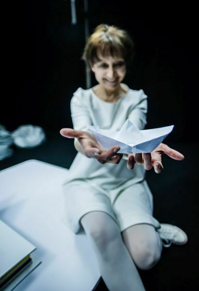 Zdjęcie ze spektaklu Książeczka. Na zdjęciu uśmiechnięta, ubrana na biało, aktorka trzyma na wyciągniętych rękach statek z papieru.