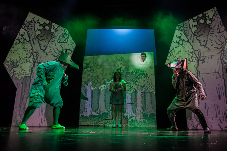 Zdjęcie ze spektaklu Maleńkie krolestwo Królewny Aurelki. Na scenie jest ciemny las. Na przeciwko siebie stoi zielony smok i szary wilk. Przygląda się im dziewczynka w krótkich spodenkach na szelkach.