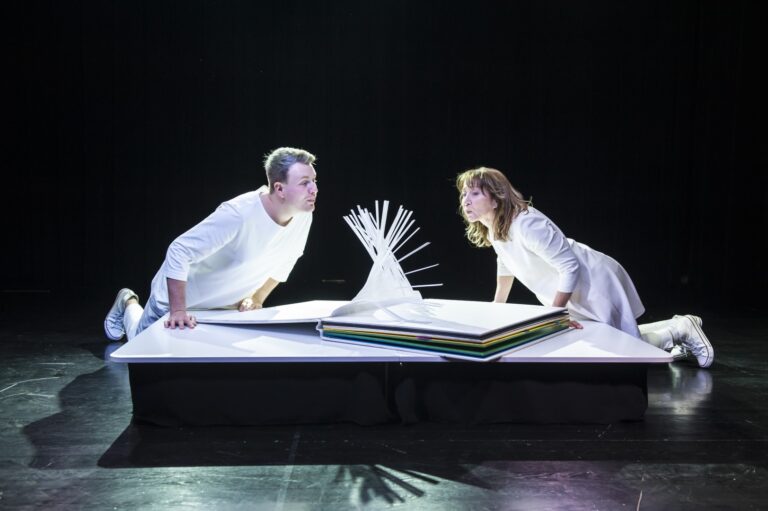Zdjęcie przedstawia dwie postacie ubrane na biało zwrócone do siebie. Pochylają się nad wielką książką, z której wyłaniają się ruchome elementy, które aktorzy pobudzają poprzez podmuch. Zdjęcie stanowi zapis ze sceny spektaklu pt. "Książeczka"