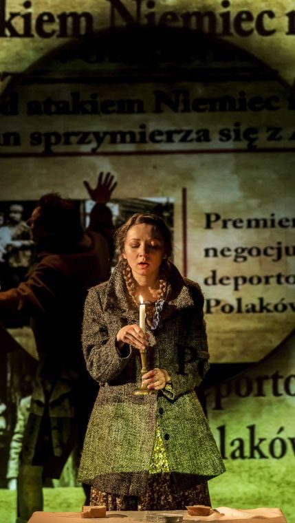 Zdjęcie ze spektaklu Jeśli nie powiesz, kto będzie wiedział? Dziewczynka w warkoczykach trzyma w dłoniach długą świeczkę. W tle widać powiększone fragmenty gazet dotyczące deportacji Polaków.