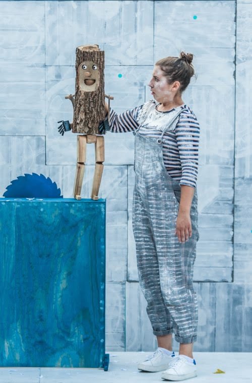 Zdjęcie ze spektaklu Pinokio. Na niebieskim stoliku z wystającą piłą do drewna stoi drewniana lalka. Trzyma ją ubrana w ogrodniczki i bluzkę w paski aktorka.