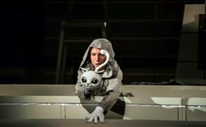 Obejrzyj zdjęcie w powiększeniu -  Zdjęcie ze spektaklu Przygody kota Filemona. Biały kot Filemon patrzy przed siebie. Za nim widać zdziwionego, animującego go aktora w jasnym kapturze.