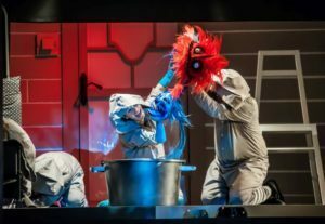 Obejrzyj zdjęcie w powiększeniu -  Zdjęcie ze spektaklu Przygody kota Filemona. Nad dużym garnkiem widać dwie lalki - niebieskiego Wodnika i czerwonego Ognika. Lalki trzymają animujący je aktorzy.