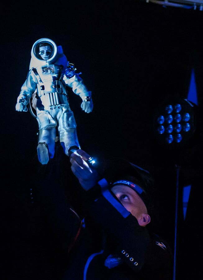 Zdjęcie ze spektaklu Ile waży jabłko na Marsie? Astronauta w ciemnym stroju patrzy w górę na trzymaną w ręku lalkę austronauty w białym, grubym kombinezonie.
