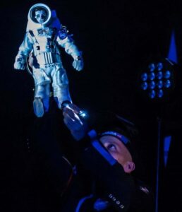 Obejrzyj zdjęcie w powiększeniu -  Zdjęcie ze spektaklu Ile waży jabło na Marsie? Kosmonauta podnosi rękę do góry - w ręce trzyma lalkę ubraną w biały skafander kosmiczny.