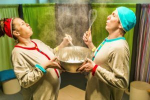 Obejrzyj zdjęcie w powiększeniu -  Photo from the play "A Kitchen Full of Surprises". The photo shows two actors (a man and a woman) in pajamas holding a large bowl of flour. Flour is also sprayed in the air and the actors blow into it.