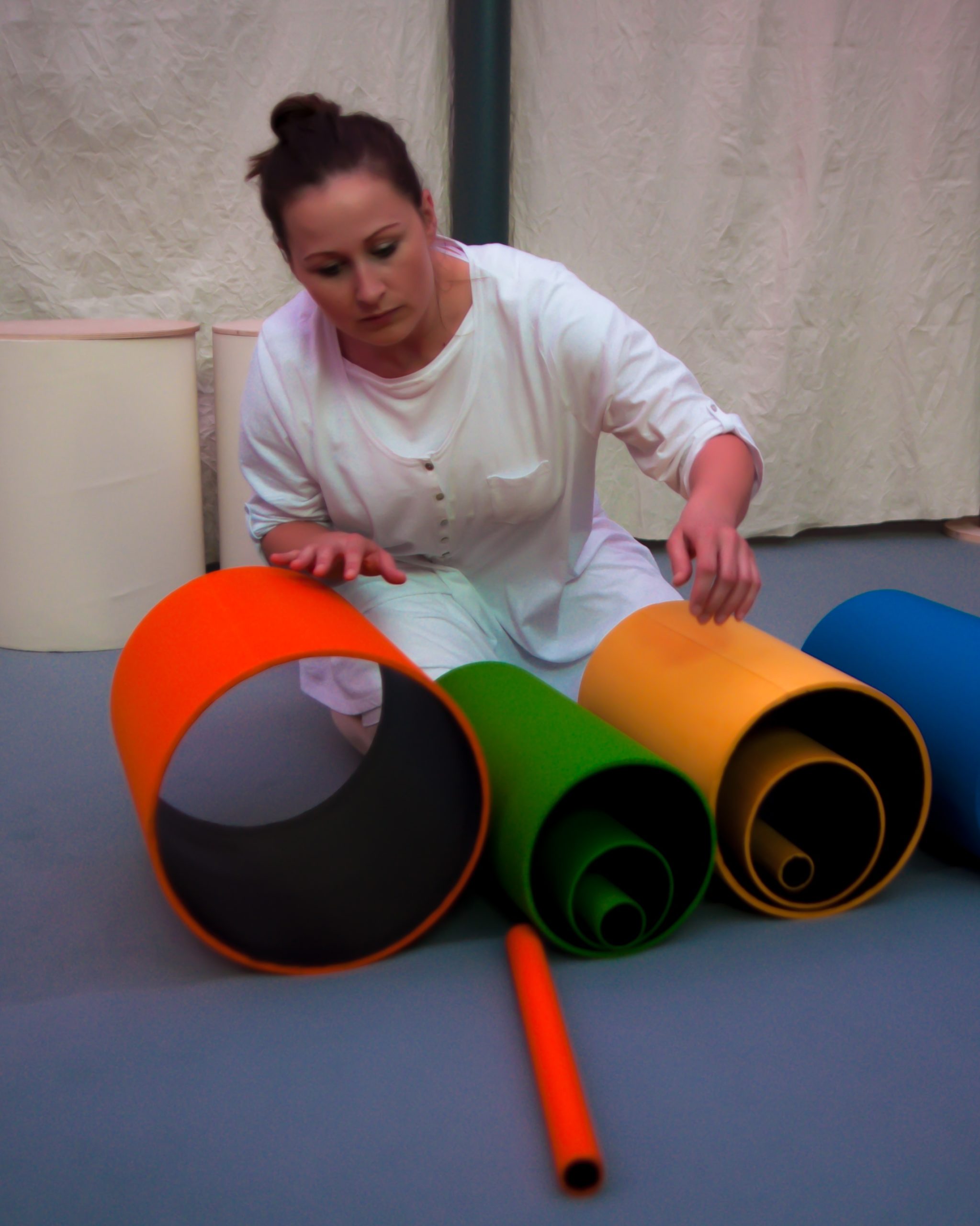 Na zdjęciu widać aktorkę kucającą nad kolorowymi tubami o różnych wielkościach, ułożonymi w jednej lini. 