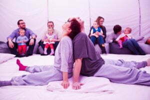 Obejrzyj zdjęcie w powiększeniu -  Photo from a play "Sleep" In a small, warm space against the wall, sits an audience with young children. In the foreground, two actors, dressed in grey T-shirts and trousers, sit with their backs against each other. Look up.