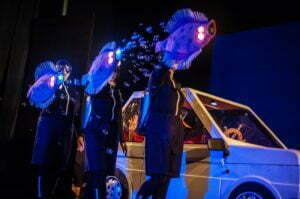 Obejrzyj zdjęcie w powiększeniu -  Photo from the show. The actors dressed in black carry 3 illuminated large Sole fish at the height of their heads. A white car stands behind them - Fiat 126 p.