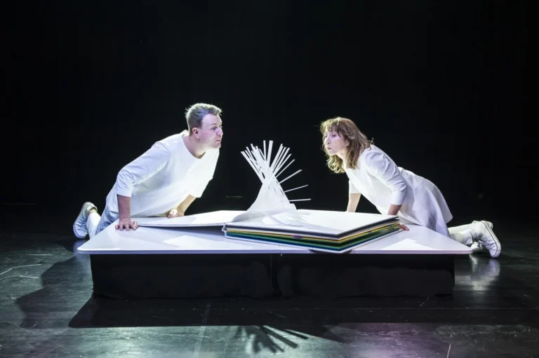 The photo shows two people dressed in white facing each other. They lean over a large book from which moving elements emerge, which the actors stimulate by blowing. The photo is a recording from the scene of the play entitled "The Little Book"