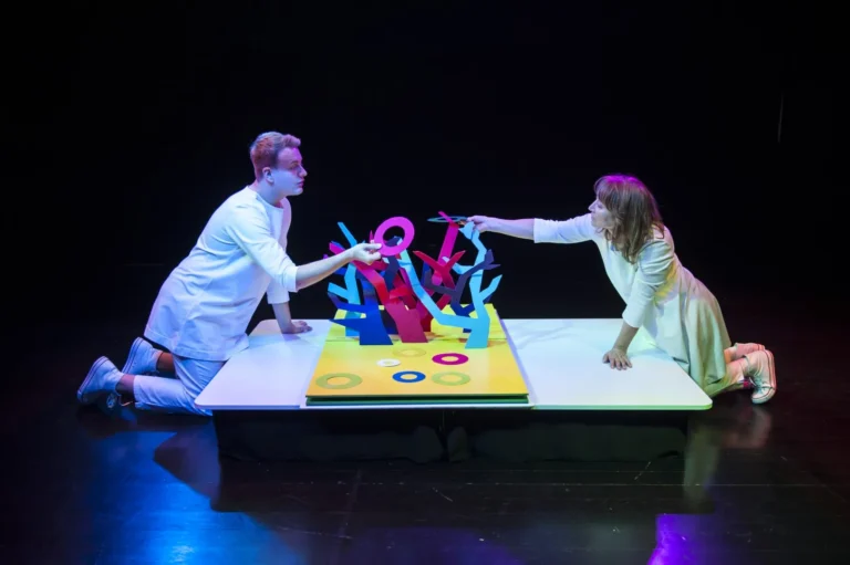 The photo shows two people dressed in white facing each other. They lean over a large book from which a colorful tree emerges and they hang colorful discs on it. The photo is a recording from the scene of the play entitled "The Little Book"