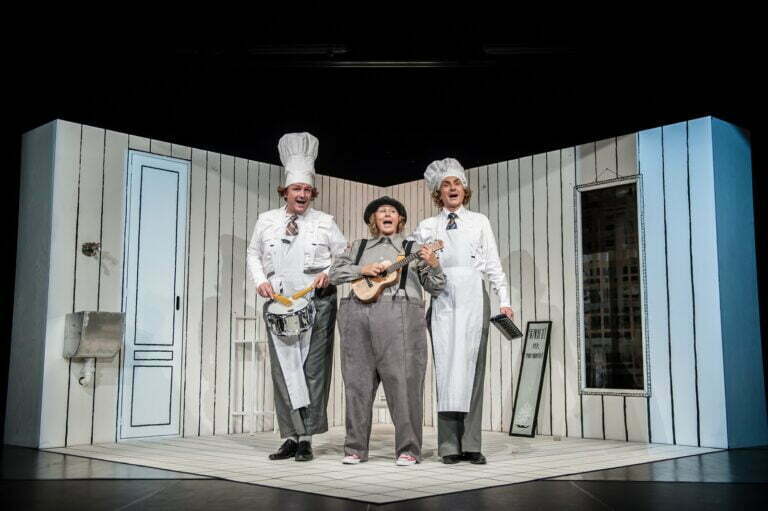 Photo from the "Spaghetti" play. Against a backdrop of white walls with black vertical stripes stand the three protagonists of the show. They sing a song. In the center stands a girl playing the ukulele, at her sides, she has two cooks - one playing the drum, the other holding a melodica.