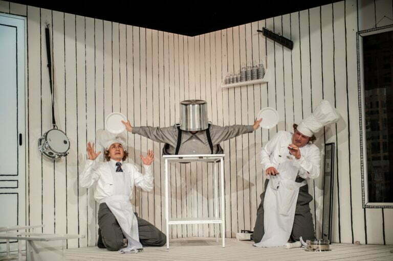 Photo from the "Spaghetti" play. On a white table lies the protagonist of the play. She has a large pot on her head and holds white plates in her outstretched hands at her sides. Underneath these plates, the chefs crouch and lean back in evasion.