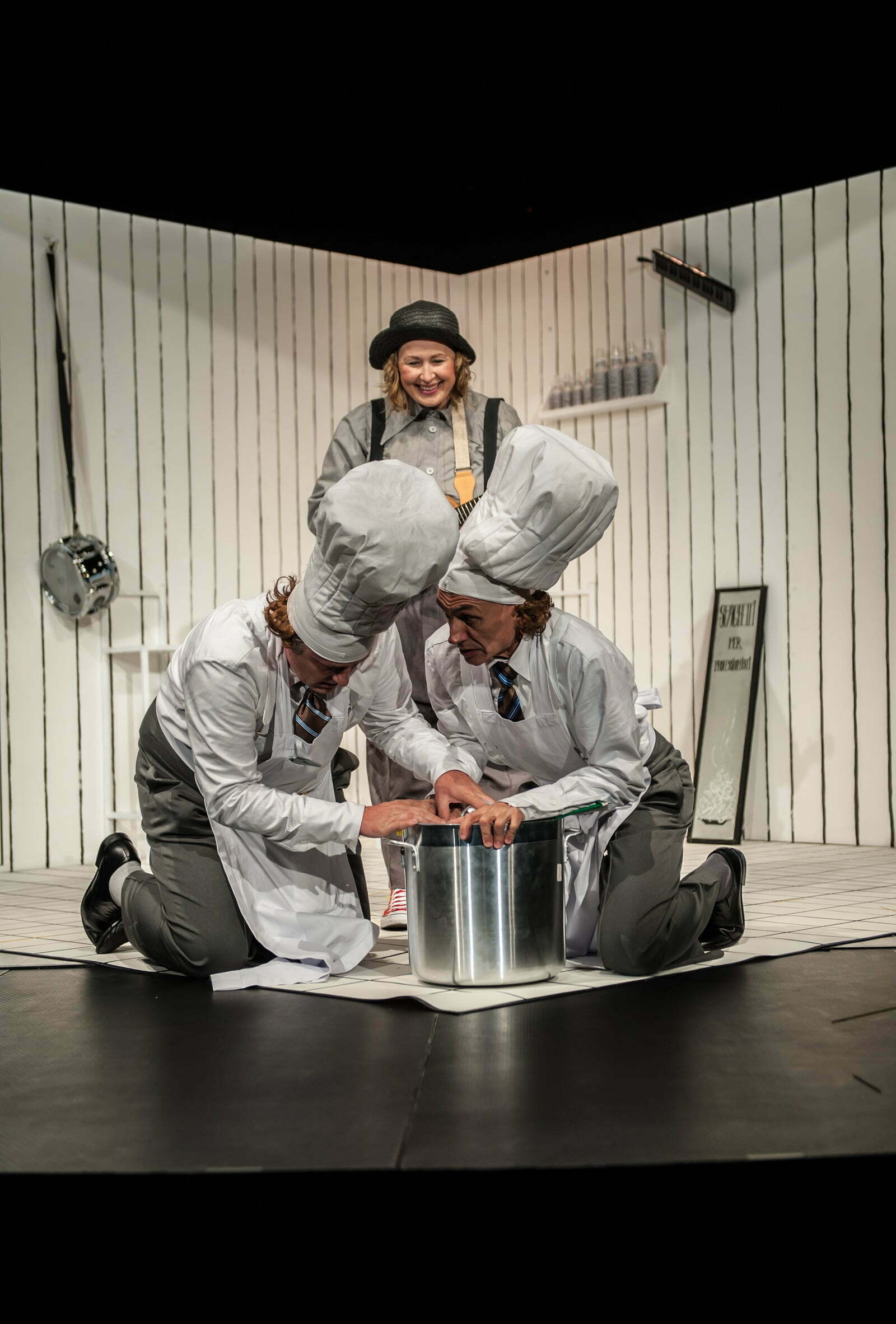 Zdjęcie ze spektaklu Spaghetti. Na pierwszym planie dwóch kucharzy pochyla się nad dużym garnkiem i przytrzymuje jego pokrywkę. Za nimi uśmiecha się inna bohaterka spektaklu.