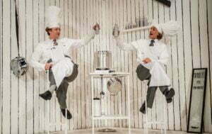 Obejrzyj zdjęcie w powiększeniu -  Photo from the "Spaghetti" play. Two chefs sit on high bar stools and lift up bottles of drink as toast. The background is white walls with black vertical stripes.