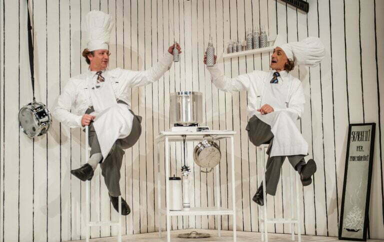 Zdjęcie ze spektaklu Spaghetti. Dwóch kucharzy siedzi na wysokich stołkach barowych i podnosi do góry buteleczki z napojem na znak toastu. Tłem są białe ściany w czarne pionowe paski.
