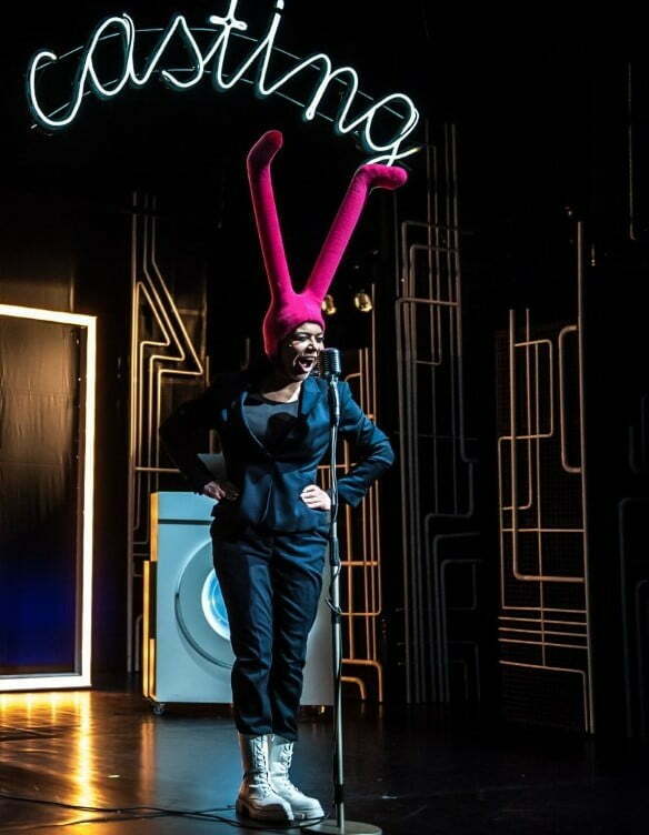 Photo from the play. A singing figure with pink tights on her head stands in front of the microphone. In the background, you can see a washing machine and lots of interconnected plumbing pipes. An inscription hangs at the top: casting.