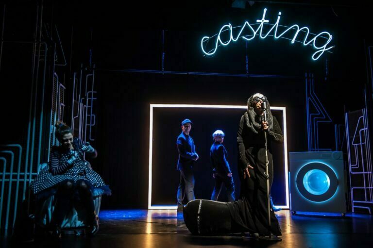 Photo from the play Black Sock with big glasses sings into the microphone. Above it a large glowing casting sign. In the background, you can see a washing machine and lots of connected plumbing pipes.