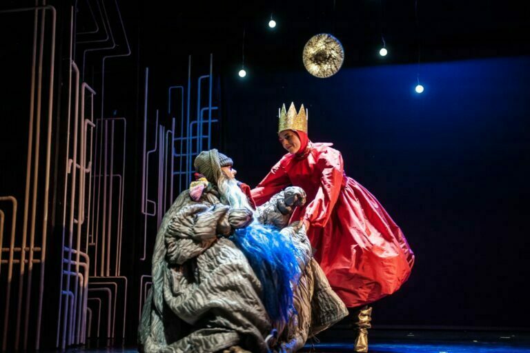 Photo from the play A homeless man with a long blue beard sits in the middle of the stage. The Queen, dressed in a red gown and golden crown, leans over him. Above them hangs the moon and stars.
