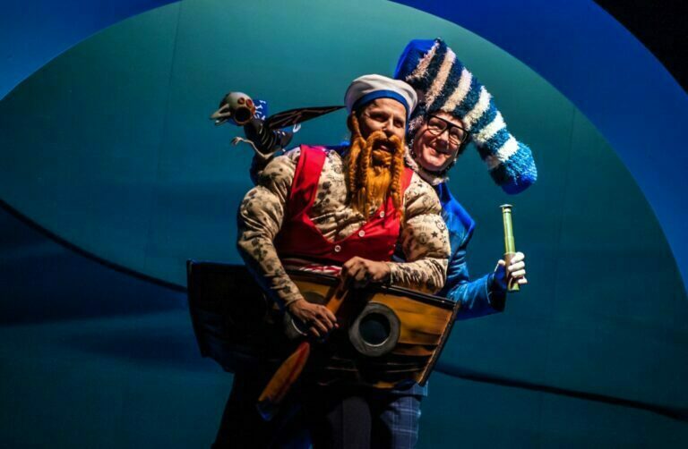 Zdjęcie ze spektaklu Niesamowite przygody Skarpetek. Marynarz z długą brodą i wąsami trzyma w ręku małe wiosło. Na jego ramieniu siedzi ptak. Zza marynarza wychyla się aktor grający Skarpetkę w paski, który trzyma w ręku lunetę.