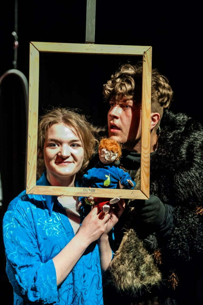 Kobieta i mężczyzna wyglądają przez drewnianą ramkę. W dłoniach trzymają lalkę przypominającą chłopca. Scena ze spektaklu Teatru Baj pt. Nieopodalja