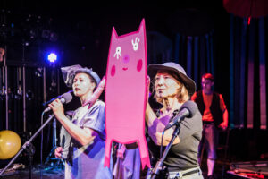 Obejrzyj zdjęcie w powiększeniu -  Photo from the concert "The Climb" Two actresses dressed in grey costumes stand in front of the microphones. One of them is holding a large, pink, cardboard cut-out pig.