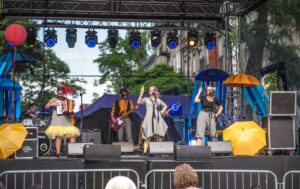 Obejrzyj zdjęcie w powiększeniu -  Zdjęcie z plenerowego spektaklu Wędrówka. Na dużej scenie na ulicy widać śpiewających i grających na instrumentach aktorów. Pomiędzy nimi leżą albo wiszą kolorowe parasole.