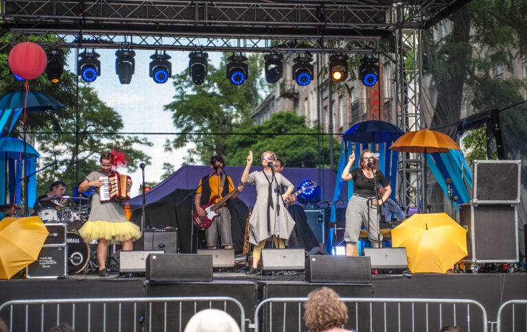 Zdjęcie z plenerowego spektaklu Wędrówka. Na dużej scenie na ulicy widać śpiewających i grających na instrumentach aktorów. Pomiędzy nimi leżą albo wiszą kolorowe parasole.
