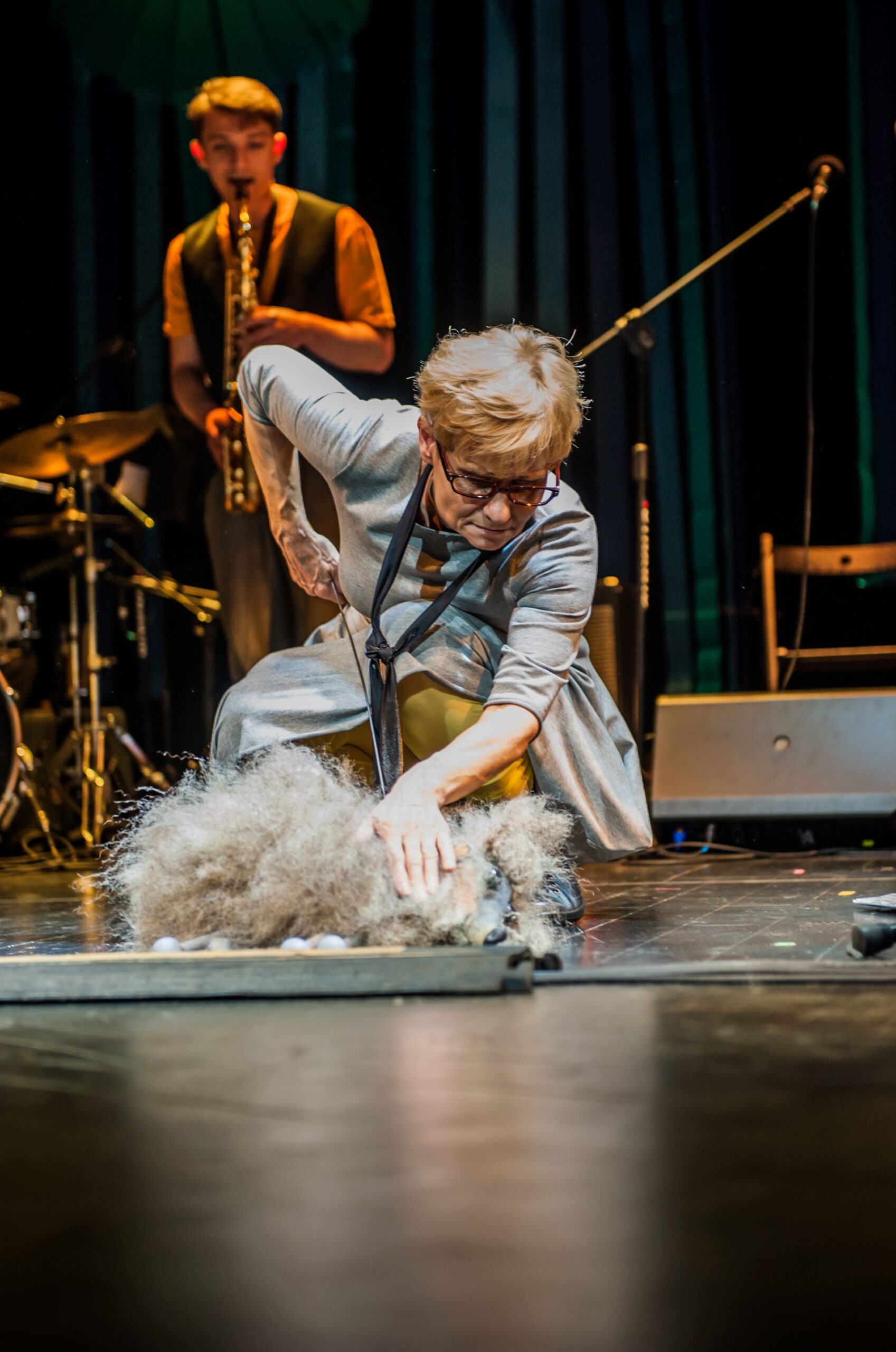 Zdjęcie ze spektaklu Wędrówka. Aktorka - blondynka w szarej sukience i okularach - schyla się nad psem pudlem. Pies jest naprawdę lalką teatralną trzymaną przez aktorkę. W tle widać grającego saksofonistę.