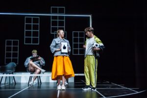 Obejrzyj zdjęcie w powiększeniu -  Pictures from the play "Peter Pan and Wendy". Two characters stand on a black stage - a boy and a girl. A girl wearing an orange skirt and a gray jacket holds a whiteboard in front of her with a graphic of a black thumb raised - a like symbol. A boy standing nearby in a green tracksuit with a ball under his arm looks at this graphic. White window frames can be seen in the background.