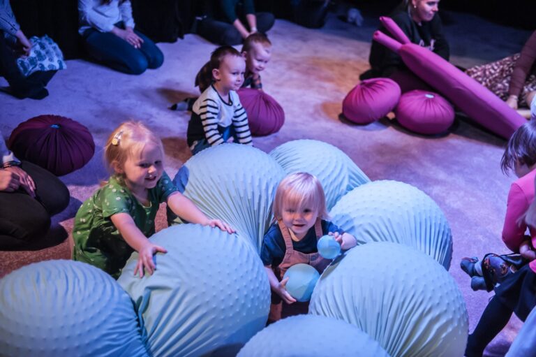 A group of young children together with actors during a play after the performance titled "The play. BODYPLAYING In the photo, the scenic elements: the fuchsia round pieces are the red blood cells in the plays and the long gray-blue connected spheres are the intestines. The performance is a journey into one's own body
