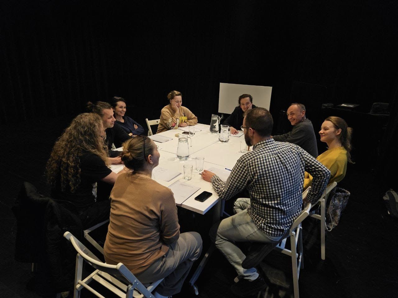Aktorzy Teatru Baj zebrani przy dużym białym stole omawiają wraz z reżyserem, producentem i inspicjentką przygotowania do najnowszej premiery pt. Czerwony Kapturek. Trwa pierwsza próba czytana.